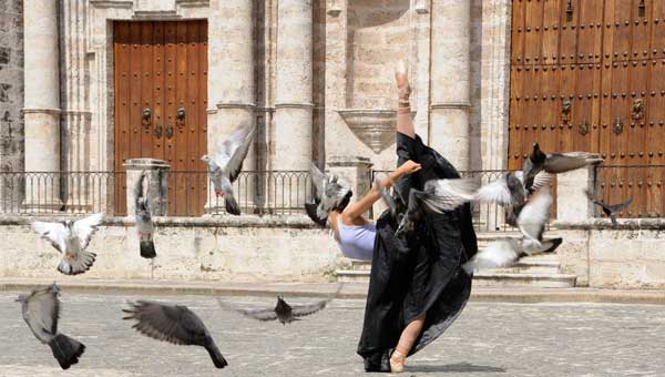 Bailarina de ballet ejecuta movimiento ante la Catedral y rodeada de palomas volando, una le cubre la cara